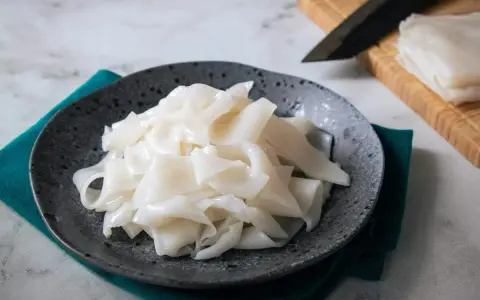 Gotowy do przygotowania makaron ryżowy na talerzu. Obok deska z nożem i stos świeżego makaronu.