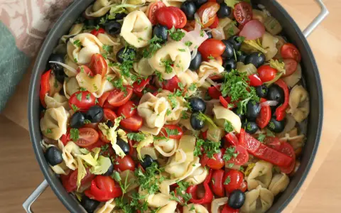 Kolorowa sałatka z tortellini, pomidorkami koktajlowymi, czarnymi oliwkami, papryką i natką pietruszki w dużej patelni.
