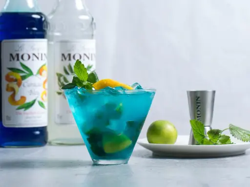 Orzeźwiający drink z syropem blue curacao smak, ozdobiony miętą i cytryną. Obok butelki syropu Monin.