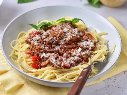 Pyszne spaghetti z bogatym sosem mięsnym na bazie passaty, posypane parmezanem i udekorowane listkami bazylii.