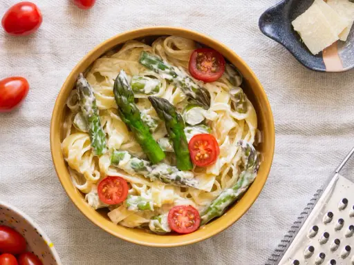 Pyszny makaron ze szparagami w kremowym sosie, ozdobiony pomidorkami koktajlowymi. Obok miseczka z parmezanem i tarką.