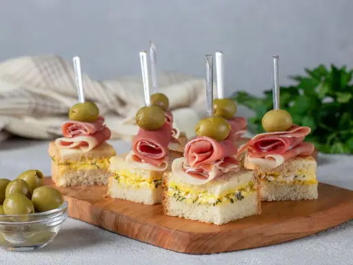 Eleganckie mini przekąski na imprezę: kanapki z szynką, jajkiem i oliwkami na drewnianej desce.