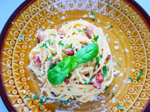Pyszne spaghetti carbonara ze śmietaną, posypane parmezanem i natką pietruszki, ozdobione listkiem bazylii.