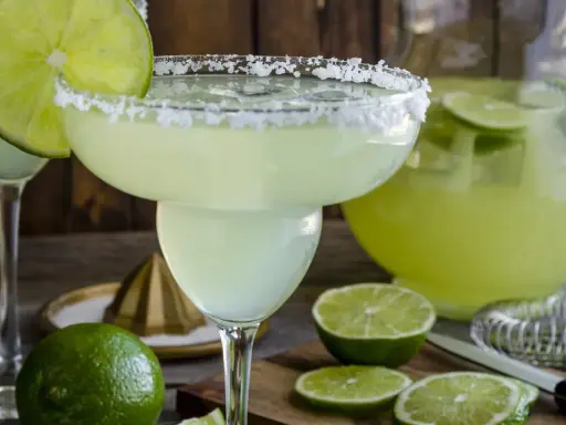 Orzeźwiająca margarita z solą na brzegu kieliszka, ozdobiona plasterkiem limonki. Obok dzbanek z drinkiem i świeże limonki.