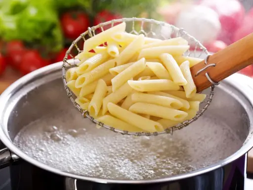 Penne makaron w cedzaku nad garnkiem z wrzącą wodą. Czas, ile się gotuje makaron, zależy od jego rodzaju.