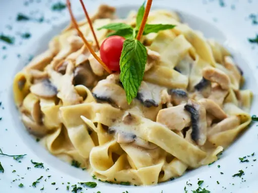 Pyszny makaron tagliatelle z kremowym sosem pieczarkowym, udekorowany pomidorkiem koktajlowym i listkiem mięty.