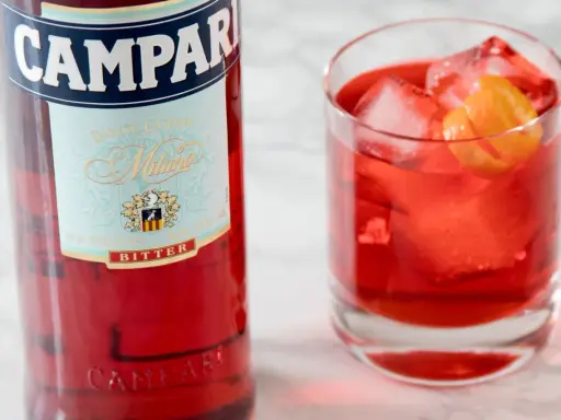 Butelka Campari i szklanka z czerwonym drinkiem, lodem i skórką pomarańczy. Idealny aperitif, który może być bazą do drinków z Aperolem.
