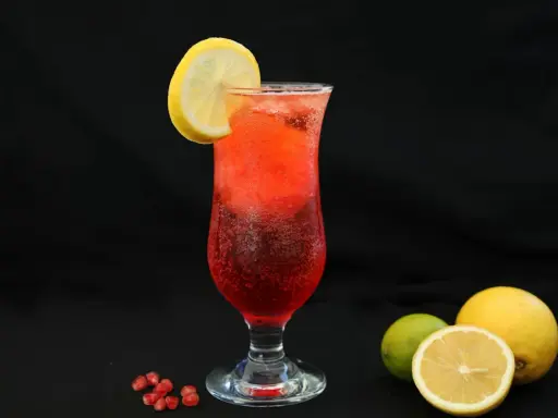 Orzeźwiający drink z granatem, ozdobiony plasterkiem cytryny. Obok leżą świeże owoce: cytryna i limonka.