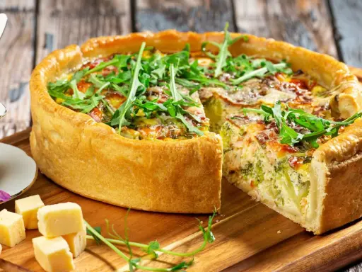 Pyszna tarta z brokułami i łososiem, udekorowana rukolą. Obok kawałki sera i sos.