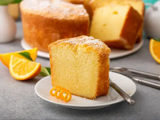 Puszysta babka pomarańczowa, oprószona cukrem pudrem, z plasterkami pomarańczy i ozdobną skórką na talerzyku.
