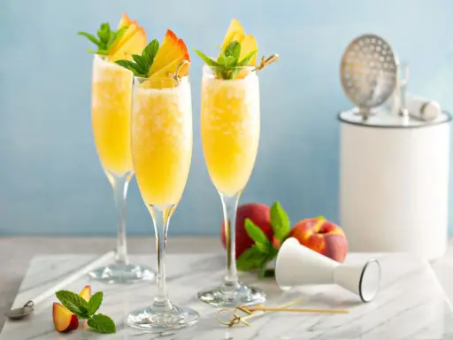 Trzy orzeźwiające drinki mimoza w kieliszkach flute, ozdobione brzoskwinią i miętą, na marmurowym blacie.
