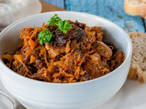 Pyszny bigos w białej misce, z gałązką natki pietruszki. Obok kromki chleba. Idealny na zimowe wieczory.