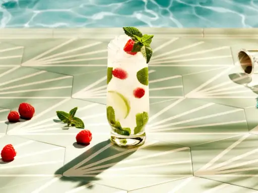 Orzeźwiający drink z malinami i miętą, idealny do Bacardi malinowego. Obok leżą maliny, a w tle widać basen i okulary przeciwsłoneczne.