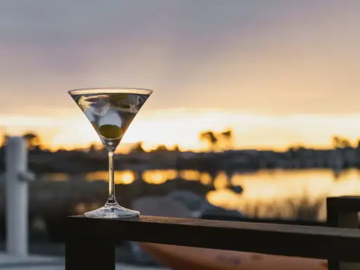 Idealny martini wstrząśnięte nie mieszane, z oliwką, na tle zachodzącego słońca nad wodą.