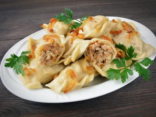 Pyszne pierogi z mięsem i kapustą, okraszone skwarkami i udekorowane natką pietruszki, na białym talerzu.