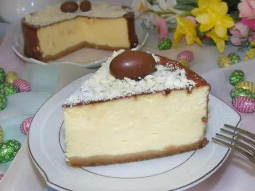 Pyszny sernik z mascarpone, udekorowany czekoladowymi jajeczkami i wiórkami białej czekolady, na tle wielkanocnych ozdób.