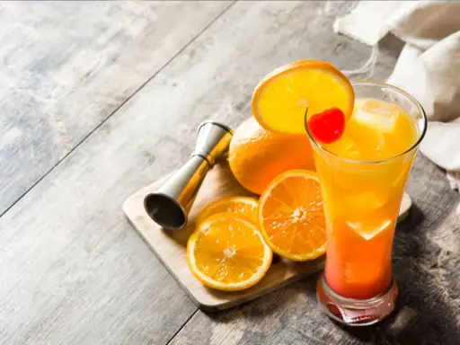 Kolorowy drink tequila sunrise z pomarańczą i wisienką, obok pomarańcze i miarka barmańska.