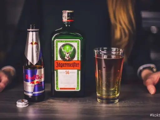 Jägermeister z czym pić? Klasycznie z Red Bullem, tworząc popularny "Jägerbomb". Na stole butelka Jägermeistera, Red Bull i szklanka z drinkiem.