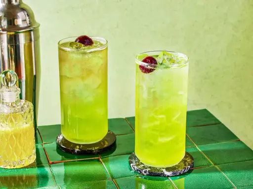 Dwa orzeźwiające drinki Tokyo Ice Tea z wiśniami i lodem, obok shakera i buteleczki z likierem.