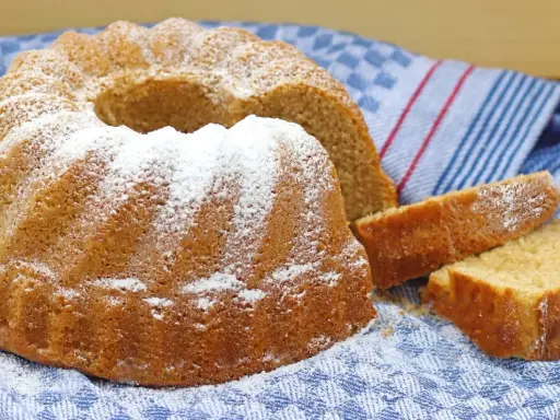 Puszysta babka wielkanocna, oprószona cukrem pudrem, leży na niebiesko-białym ręczniku. Obok niej kilka kromek ciasta.
