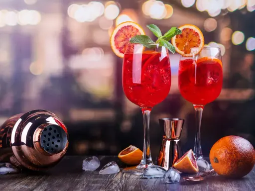 Dwa kieliszki orzeźwiającego drinka z prosecco z czymś czerwonym, plasterkami pomarańczy i miętą. Obok shaker i pomarańcze.