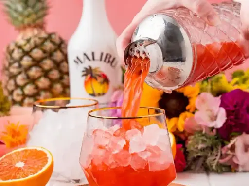 Malibu z sokiem ananasowym nalewany do szklanki z lodem. W tle ananas, butelka Malibu i kwiaty.