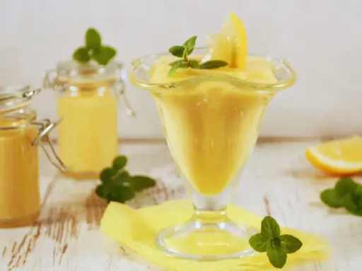 Aksamitny lemon curd w pucharku, ozdobiony plasterkiem cytryny i miętą. Obok słoiczki z tym samym kremowym przysmakiem.