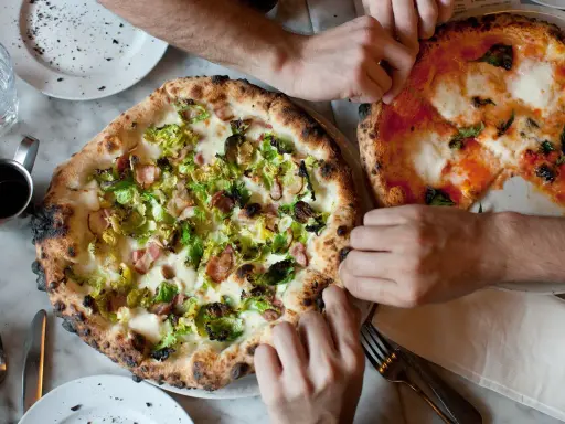 Dwie pizze na stole, jedna z brukselką i boczkiem, druga margherita. Ludzie sięgają po kawałki, pokazując, ile wyrasta ciasto na pizze.