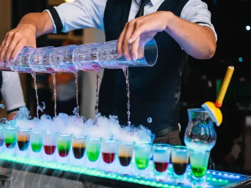 Barman nalewa kolorowe shoty z wielopoziomowego shakera. Efektowny drink bar diy z dymem i neonowym oświetleniem.