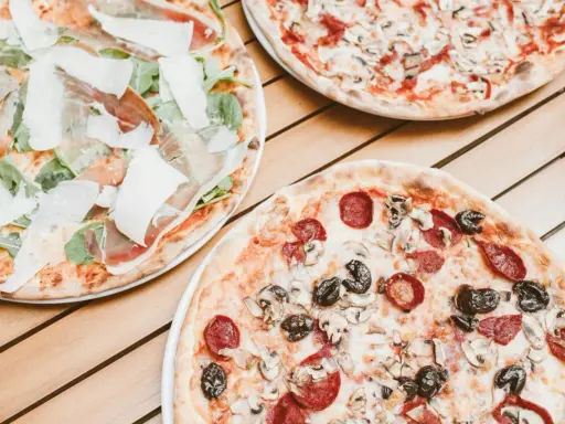 Trzy pizze na drewnianym stole. Jedna z szynką parmeńską i rukolą, druga z salami i pieczarkami, trzecia z pieczarkami. Szukasz alternatywy, jaki ser do pizzy zamiast mozzarelli?
