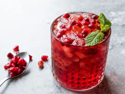 Orzeźwiający drink z grenadyną, lodem i pestkami granatu, ozdobiony listkiem mięty. Obok łyżeczka z owocem.