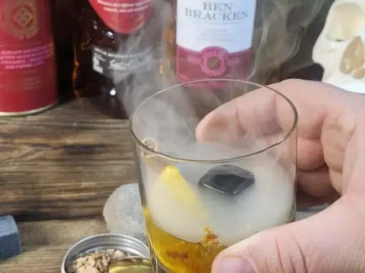 Szklanka z drinkiem otoczona dymem, obok butelki whisky i czaszka. Wyjątkowe drinki z dymem.