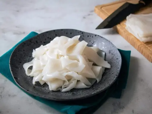 Gotowy do przygotowania makaron ryżowy na talerzu. Obok deska z nożem i stos świeżego makaronu.