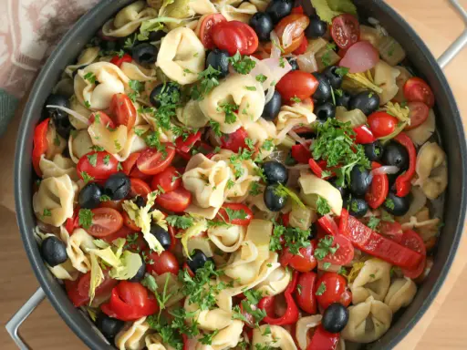 Kolorowa sałatka z tortellini, pomidorkami koktajlowymi, czarnymi oliwkami, papryką i natką pietruszki w dużej patelni.