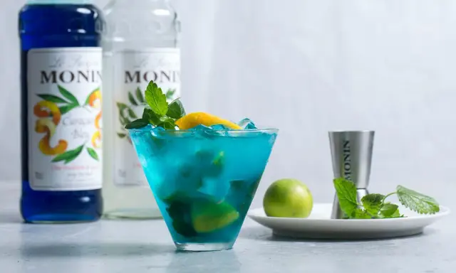 Orzeźwiający drink z syropem blue curacao smak, ozdobiony miętą i cytryną. Obok butelki syropu Monin.