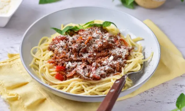 Pyszne spaghetti z bogatym sosem mięsnym na bazie passaty, posypane parmezanem i udekorowane listkami bazylii.