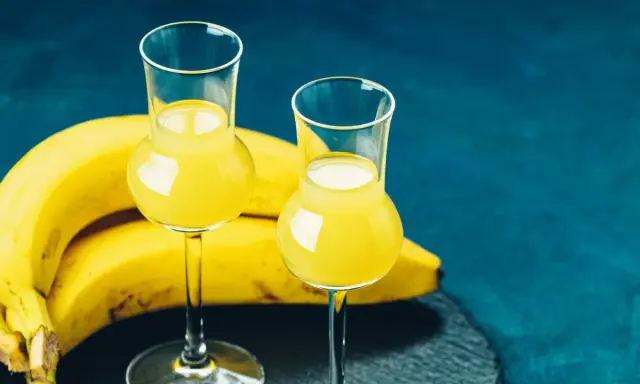Dwa kieliszki z żółtym likierem bananowym, obok kiść bananów na ciemnoniebieskim tle.