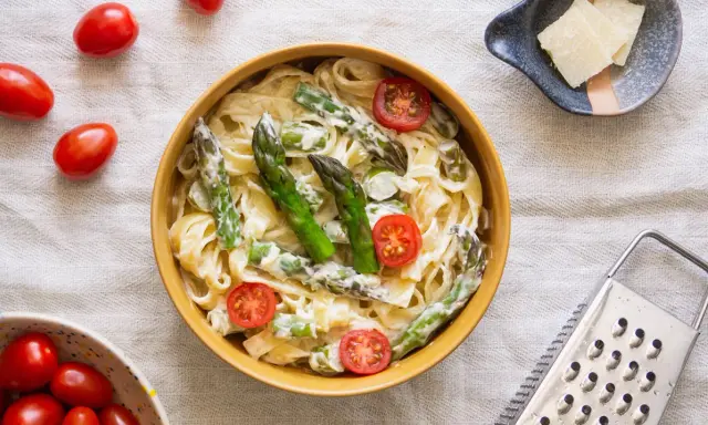 Pyszny makaron ze szparagami w kremowym sosie, ozdobiony pomidorkami koktajlowymi. Obok miseczka z parmezanem i tarką.