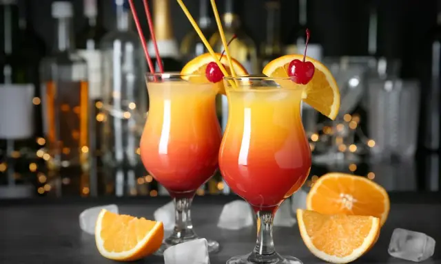 Dwa kolorowe, domowe drinki z wódką, ozdobione pomarańczą i wiśnią, z kostkami lodu i owocami w tle.