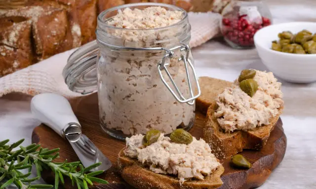 Słoik pełen pysznej pasty z tuńczyka, gotowej do rozsmarowania na chlebie. Na desce kromki z pastą i kaparami, obok gałązka rozmarynu.