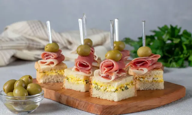 Eleganckie mini przekąski na imprezę: kanapki z szynką, jajkiem i oliwkami na drewnianej desce.