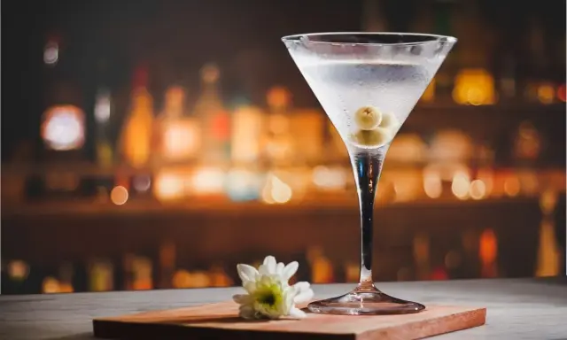Eleganckie drinki z martini, ozdobione oliwkami, stoją na blacie baru. Obok kieliszka leży biały kwiat.