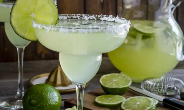 Orzeźwiająca margarita z solą na brzegu kieliszka, ozdobiona plasterkiem limonki. Obok dzbanek z drinkiem i świeże limonki.