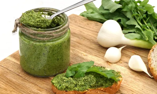 Słoik domowego pesto z rukoli, łyżka w środku, kromka chleba z pastą i świeże składniki: czosnek i rukola.