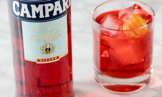 Butelka Campari i szklanka z czerwonym drinkiem, lodem i skórką pomarańczy. Idealny aperitif, który może być bazą do drinków z Aperolem.