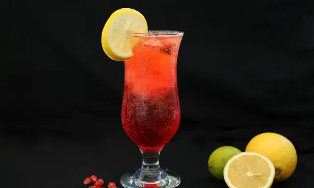 Orzeźwiający drink z granatem, ozdobiony plasterkiem cytryny. Obok leżą świeże owoce: cytryna i limonka.