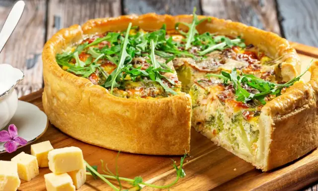 Pyszna tarta z brokułami i łososiem, udekorowana rukolą. Obok kawałki sera i sos.