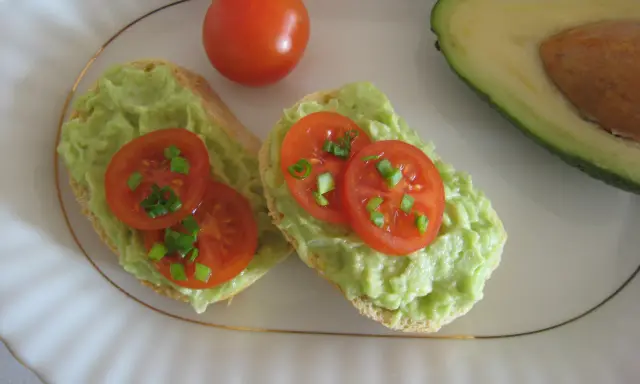 Dwa tosty z pastą z awokado, pomidorkami koktajlowymi i szczypiorkiem. Obok połówka awokado i cały pomidorek.