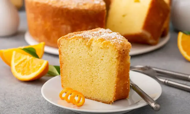 Puszysta babka pomarańczowa, oprószona cukrem pudrem, z plasterkami pomarańczy i ozdobną skórką na talerzyku.