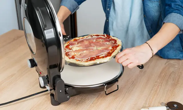 Osoba wkłada pizzę z szynką do okrągłego piekarnika. Idealny moment na pieczenie pizzy w piekarniku!